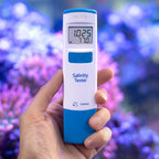 Hanna HI98319 marine salinity tester held in hand in front of a reef aquarium