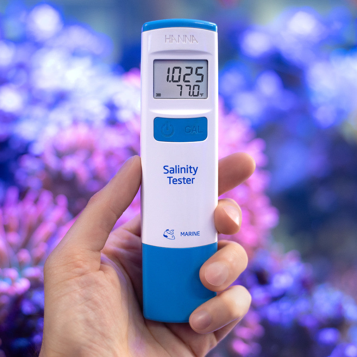 Hanna HI98319 marine salinity tester held in hand in front of a reef aquarium